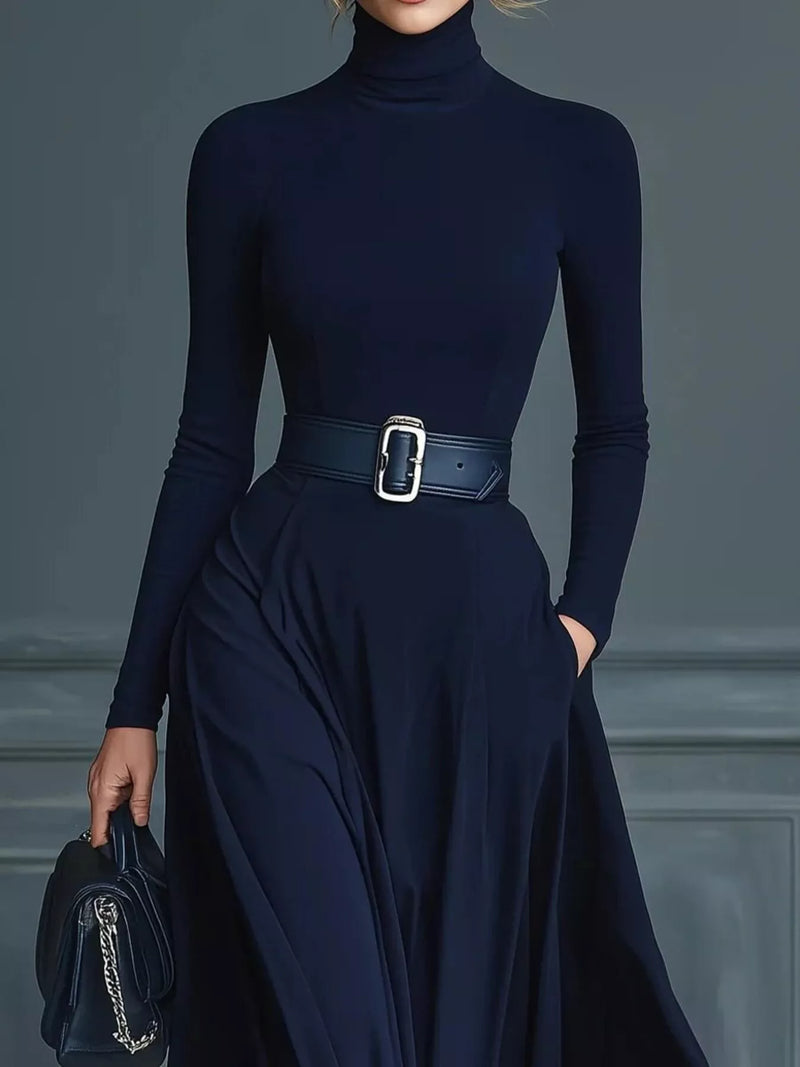 Person wearing a navy blue turtleneck and skirt with a belt, holding a matching handbag against a gray background.