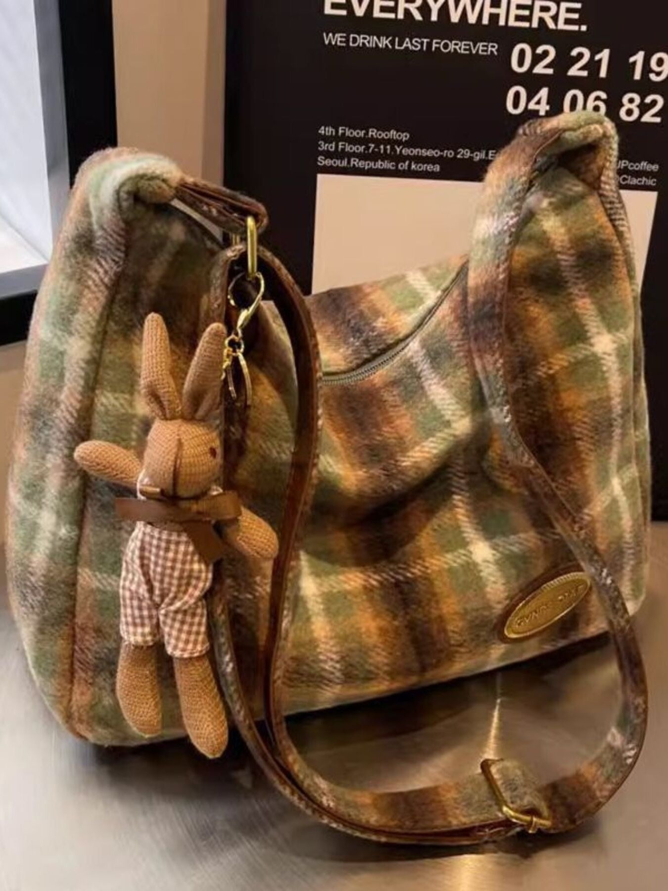 Plaid handbag with a plush bunny toy attached to the strap on a reflective surface.