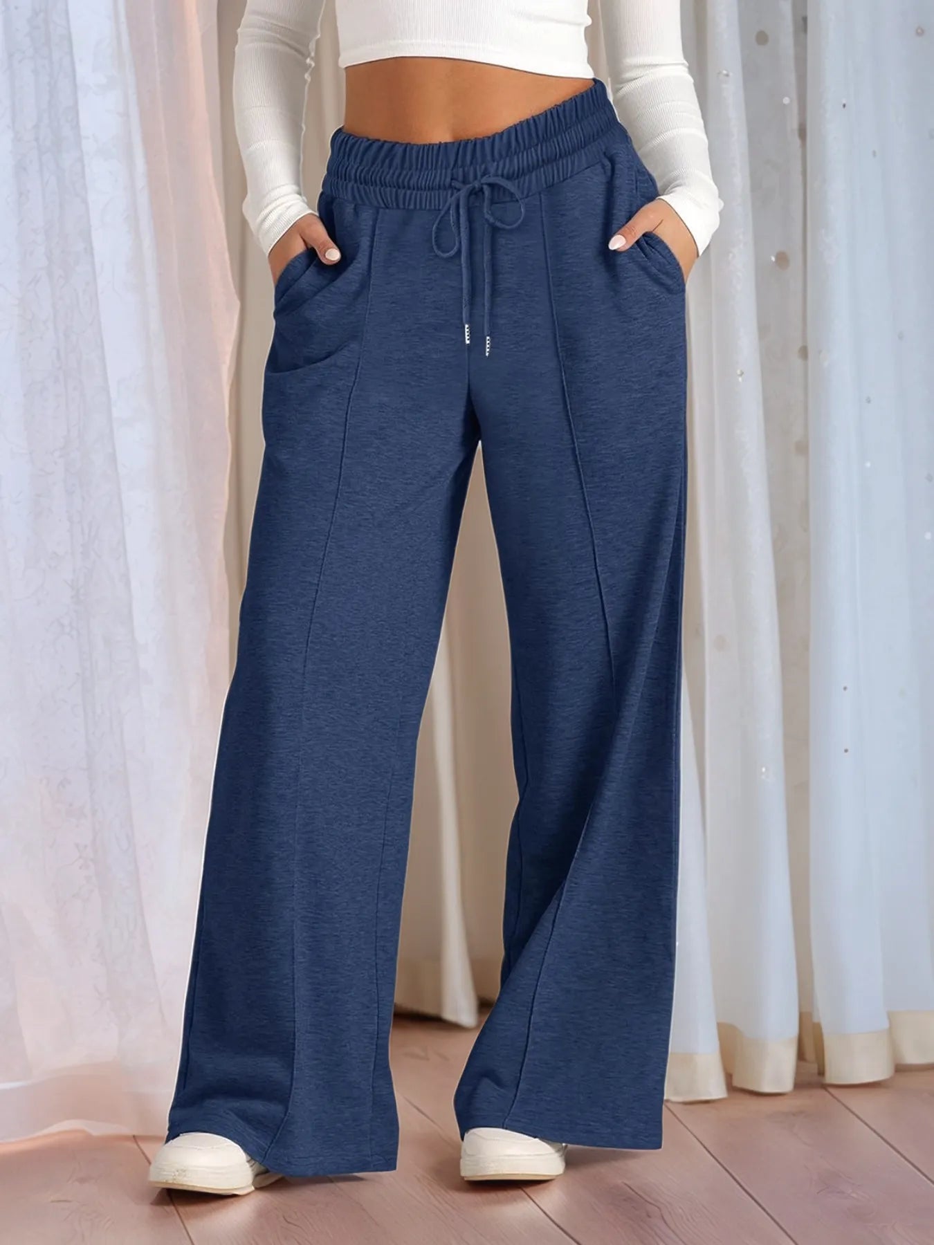 Person wearing blue wide-leg pants with a white top and shoes, standing against a light curtain background.