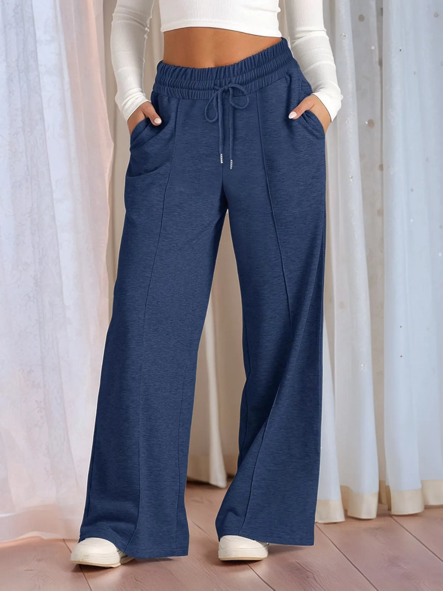 Person wearing blue wide-leg pants with a white top and shoes, standing against a light curtain background.