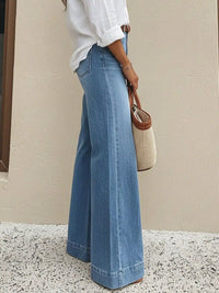 Person wearing blue wide-leg jeans and a white shirt, holding a woven bag against a beige wall.
