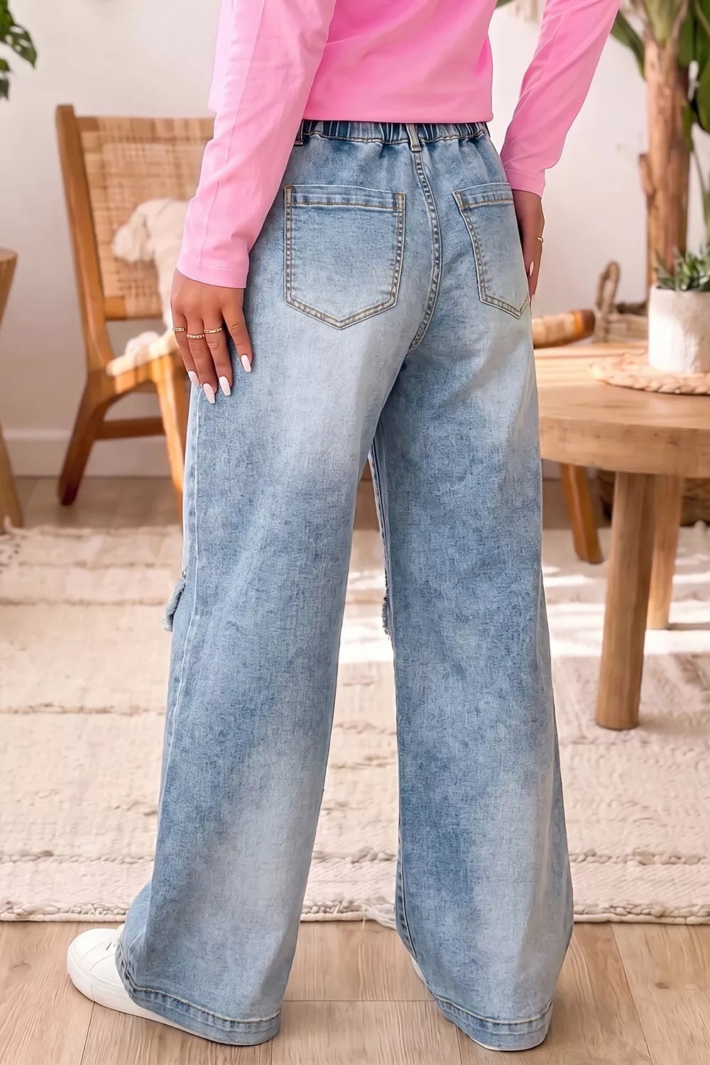 A person wearing light-wash wide-leg denim jeans and a pink long-sleeve top, shown from the back in a bright indoor room with wooden furniture and a rug.