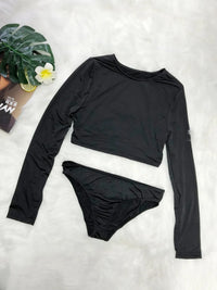 Black long-sleeve top and bikini bottom set on a white surface with a plant and magazine in the background.