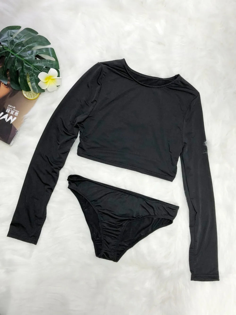 Black long-sleeve top and bikini bottom set on a white surface with a plant and magazine in the background.