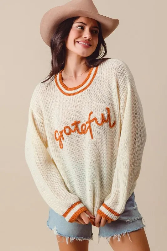 Woman wearing a cream sweater with 'grateful' text and denim shorts against a beige background