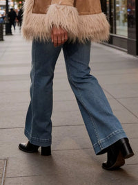 Person wearing a fur-trimmed coat, blue jeans, and black shoes on a sidewalk.