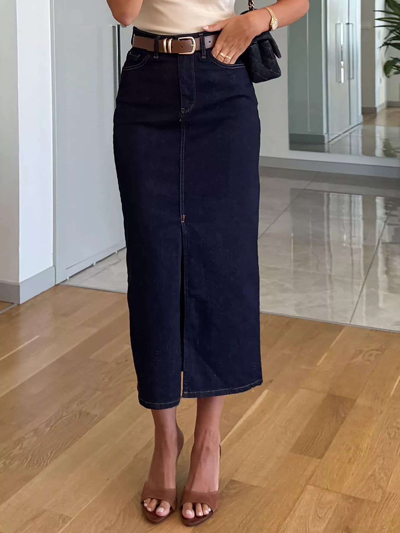Person wearing a navy denim skirt with a white top and brown sandals indoors.
