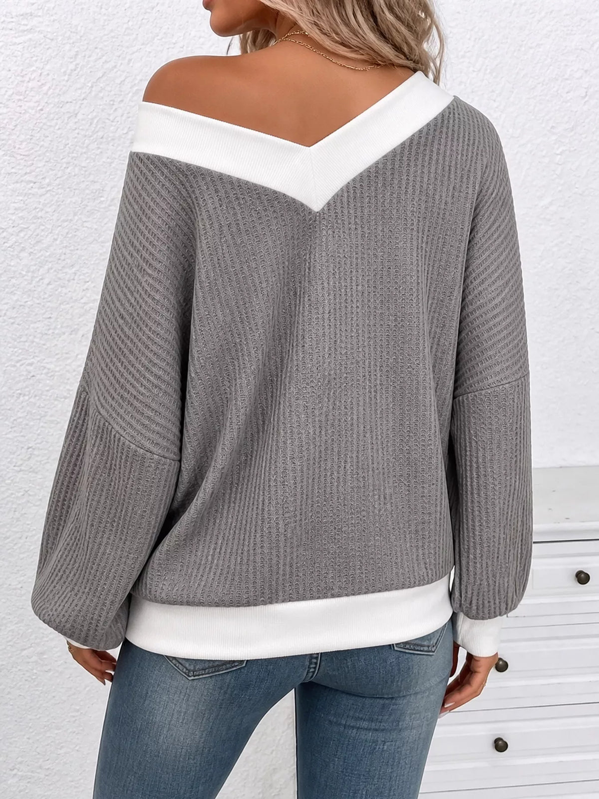 A woman wearing a gray ribbed knit sweater with contrasting white trim at the off-the-shoulder neckline, hem, and cuffs, paired with blue jeans.