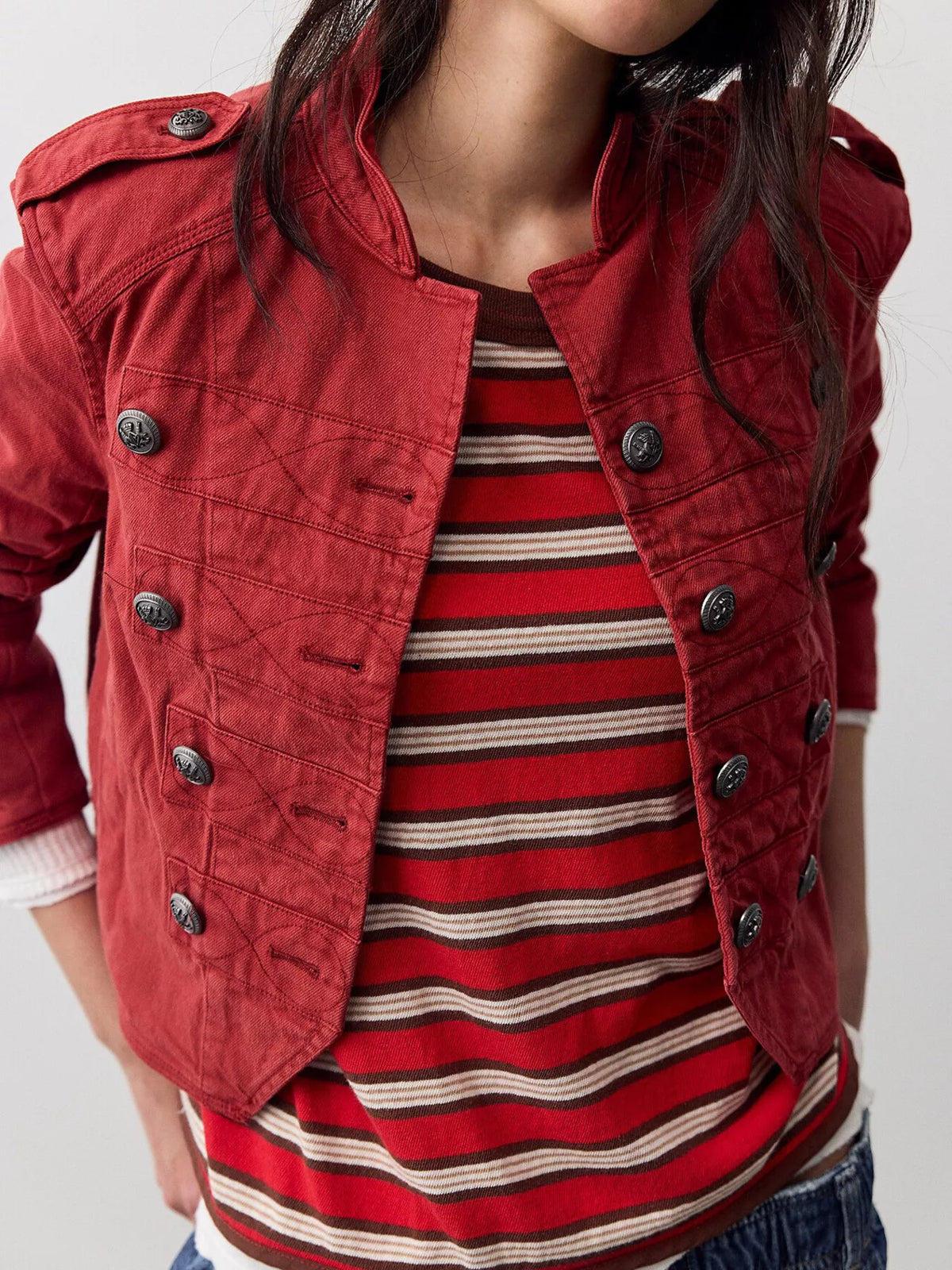 A close-up of a person wearing a red cropped jacket over a red, black, and beige striped shirt, with dark hair and jeans visible against a plain light background.