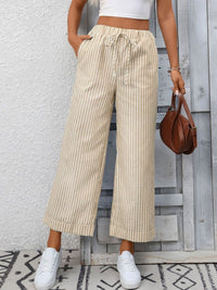 Person wearing beige striped pants and white sneakers, holding a brown handbag against a light gray wall.