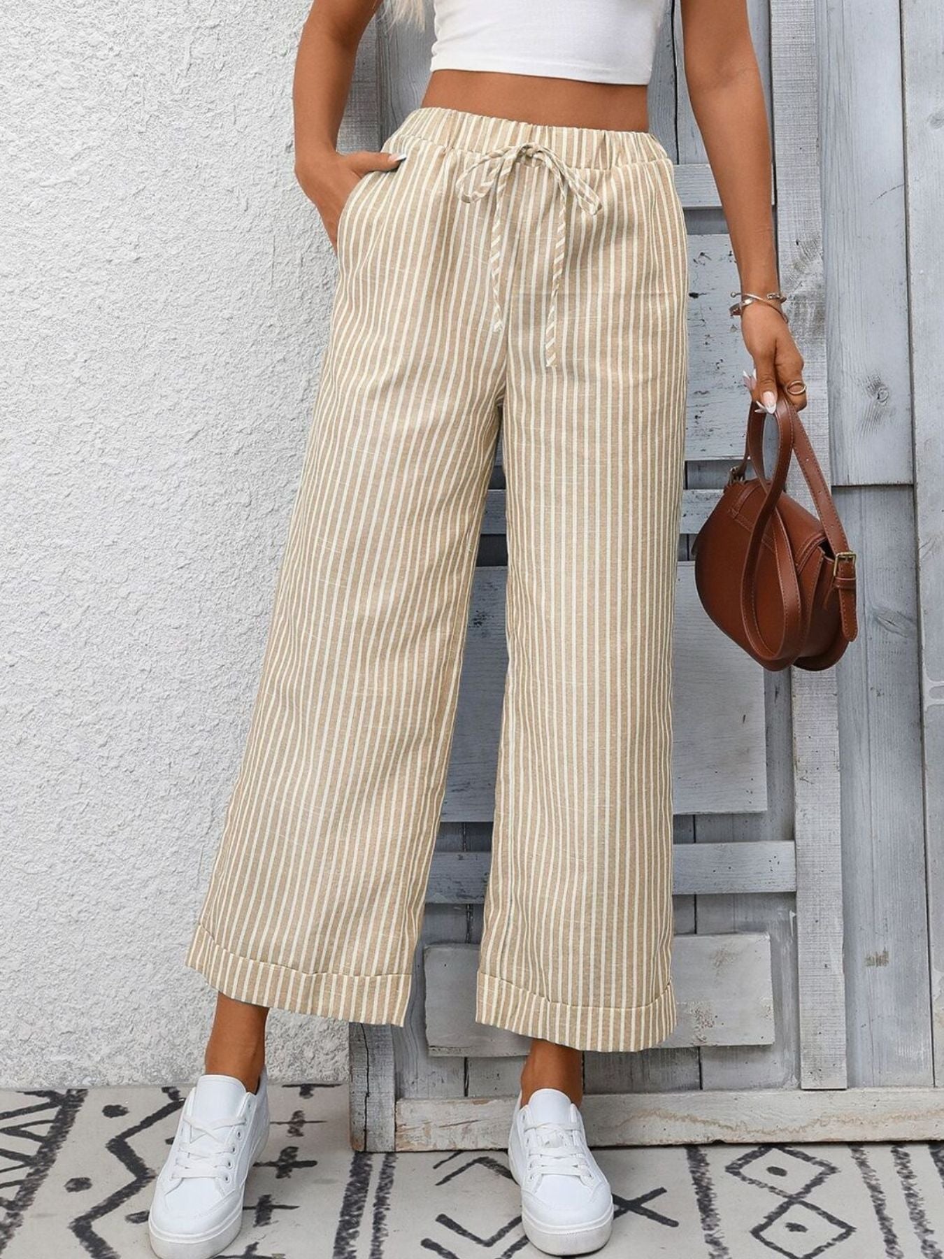 Person wearing beige striped pants and white sneakers, holding a brown handbag against a light gray wall.