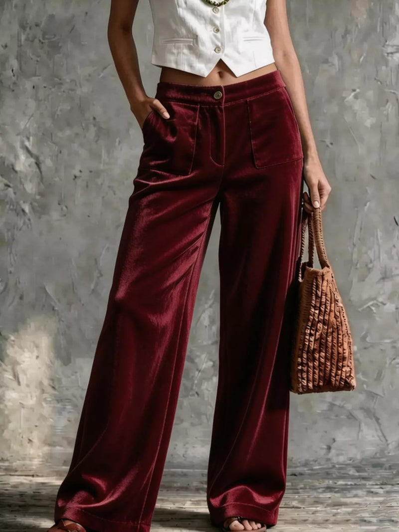 A model wearing high-waisted, wide-leg burgundy velvet trousers, styled with a white cropped vest, holding a brown woven handbag against a textured gray wall.