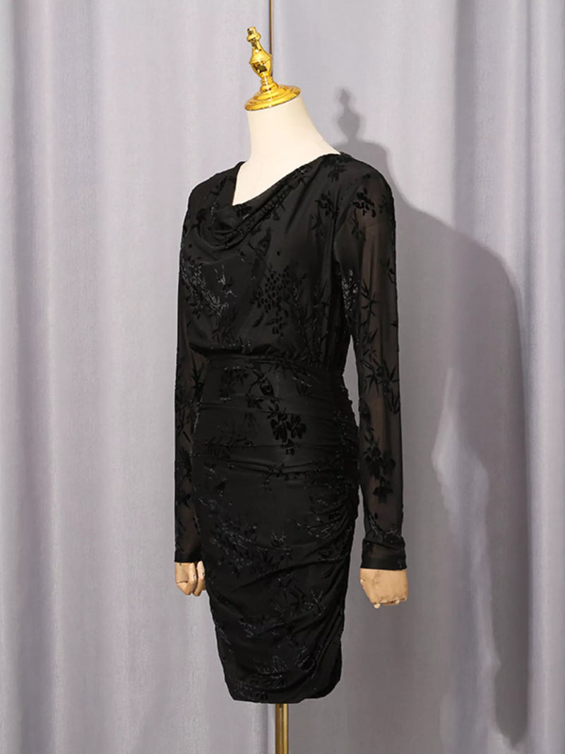 A black long-sleeve cowl-neck dress with floral lace applique, displayed on a cream mannequin with a gold neck cap against a light gray curtain backdrop.