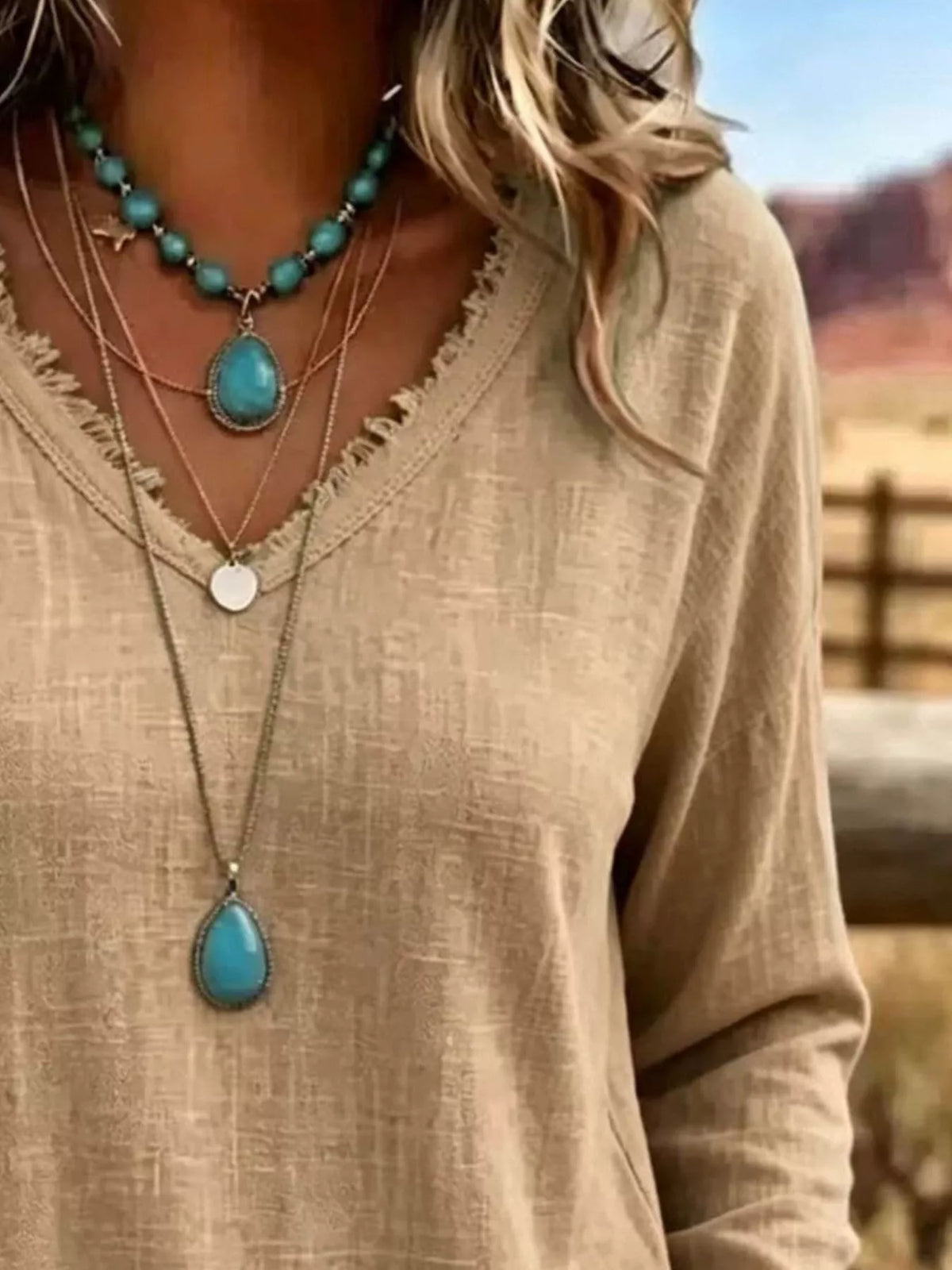 A close-up of a person wearing layered necklaces, including a turquoise beaded necklace, a turquoise teardrop pendant necklace, and a silver disc pendant necklace, paired with a beige linen top.