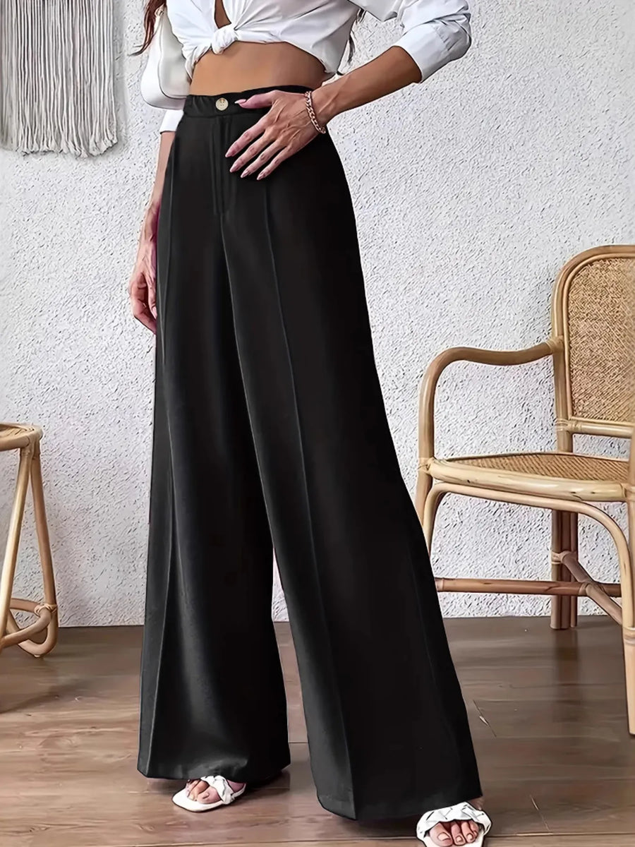 Person wearing black wide-leg pants and a white top in a room with a textured wall and wooden floor.
