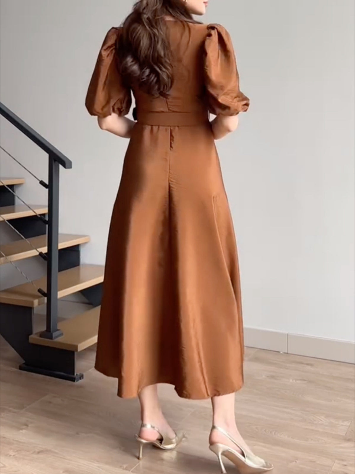 Woman wearing a brown dress standing in a room with wooden flooring and stairs.