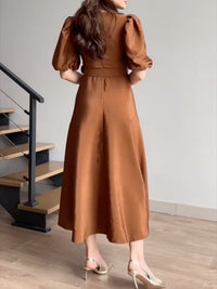 Woman wearing a brown dress standing in a room with wooden flooring and stairs.