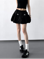 Person wearing a black skirt with white bows, black top, white socks, and black shoes on a plain background.