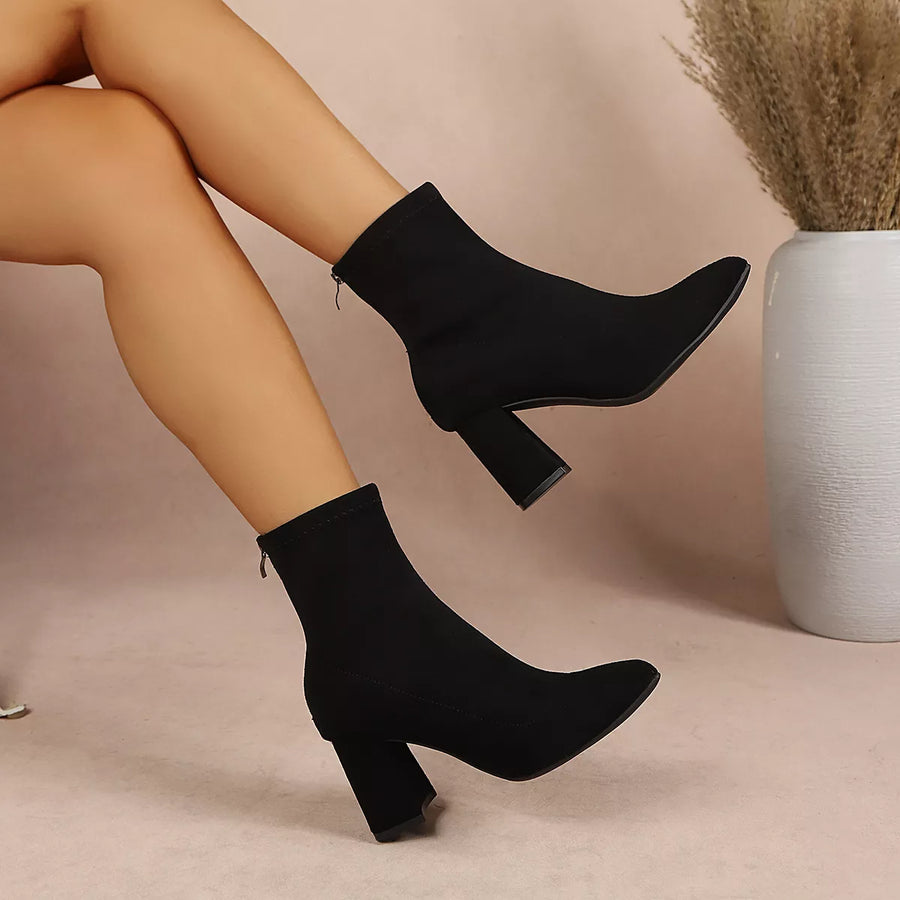 A pair of black ankle boots with block heels, shown on a model's legs against a beige background with a white vase of dried grass.