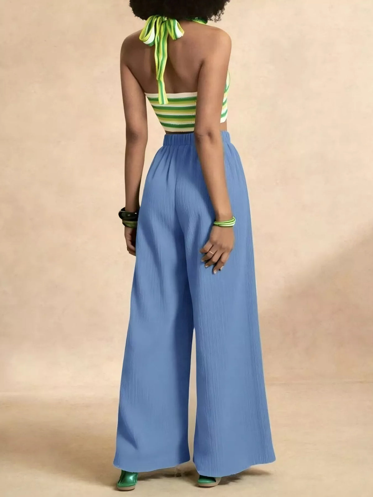 A model shown from the back wearing a green and white striped halter top with a tie-neck and high-waisted, wide-leg blue pants, set against a solid beige studio background.