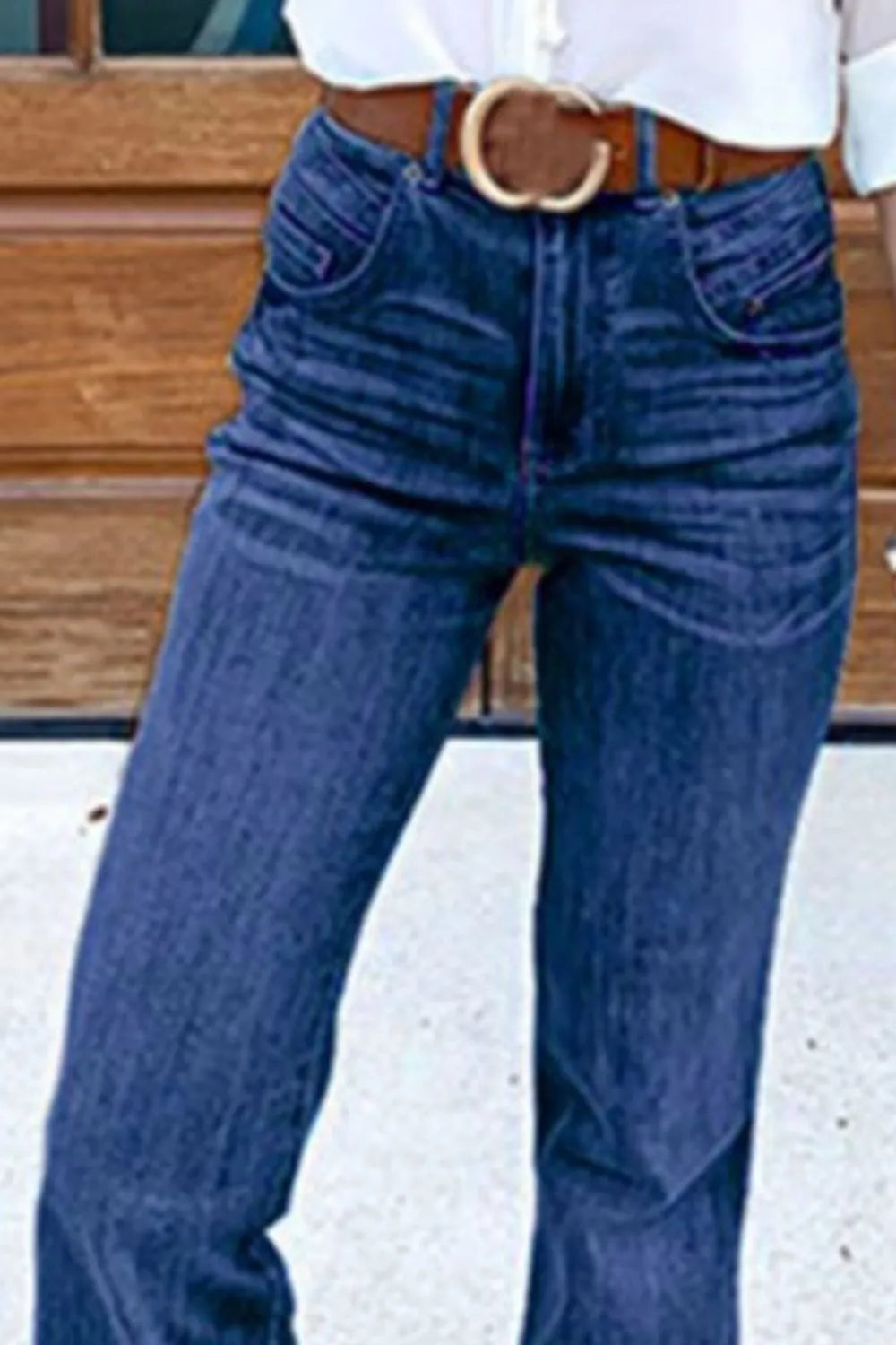 Person wearing blue jeans with a belt, standing against a wooden background.