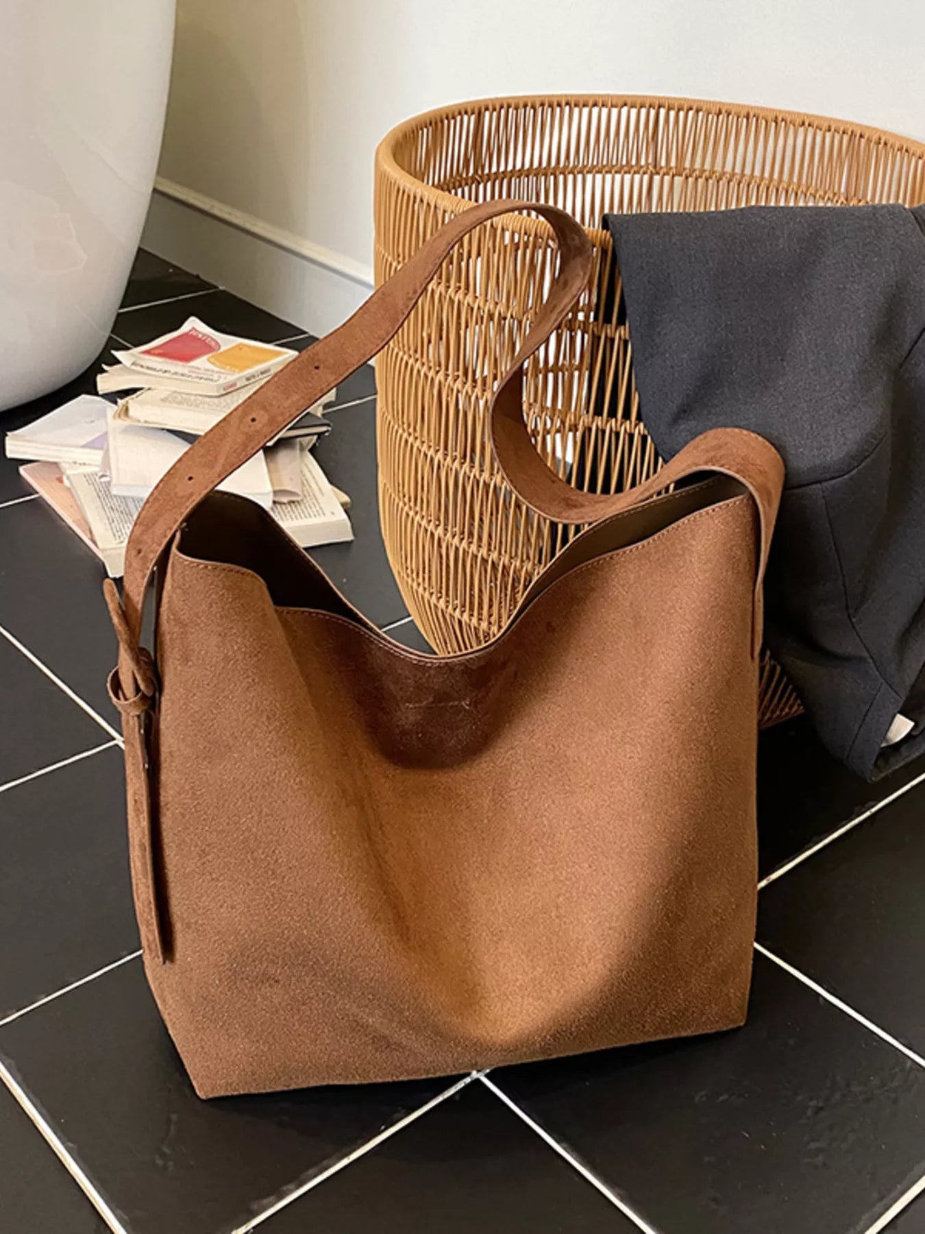 Brown leather tote bag on a black tiled floor with a wicker basket and books in the background.