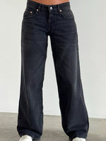 A person wearing dark washed wide-leg jeans, shown from the waist down against a plain light gray wall and concrete floor.