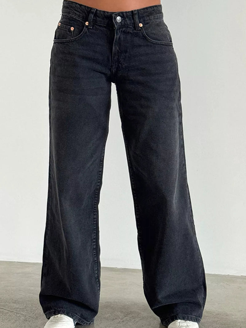 A person wearing dark washed wide-leg jeans, shown from the waist down against a plain light gray wall and concrete floor.