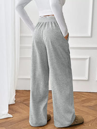 Person wearing gray sweatpants and a white long-sleeve top in a room with wooden flooring and white walls.