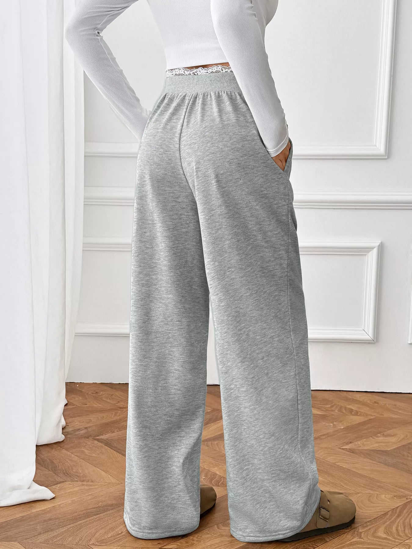 Person wearing gray sweatpants and a white long-sleeve top in a room with wooden flooring and white walls.