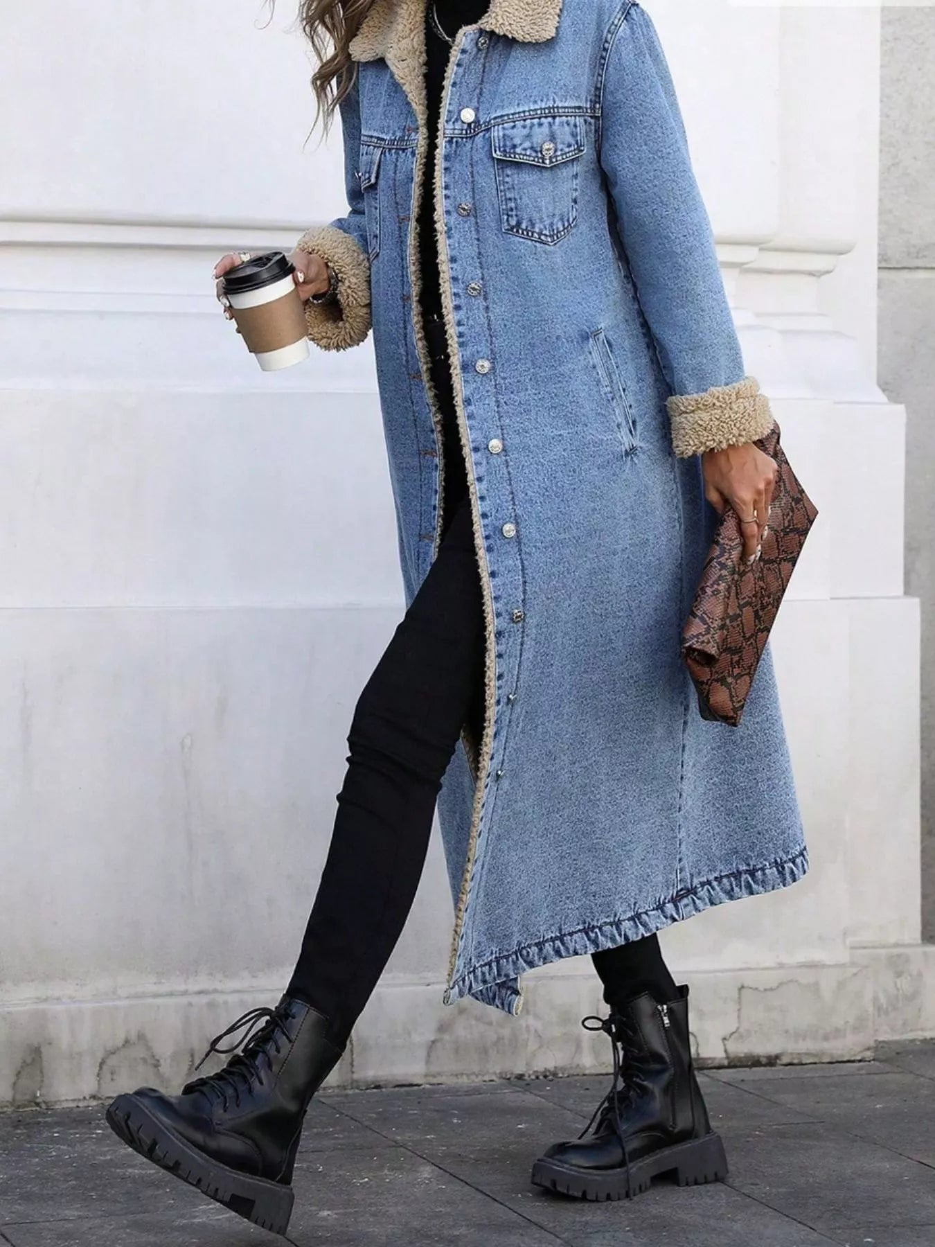 Person wearing a long denim coat with fur trim, holding a coffee cup and clutch.