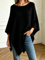 A woman wearing a black wide-sleeve poncho top with an asymmetrical hem, paired with light blue jeans, standing in a room with white walls and a wooden floor.