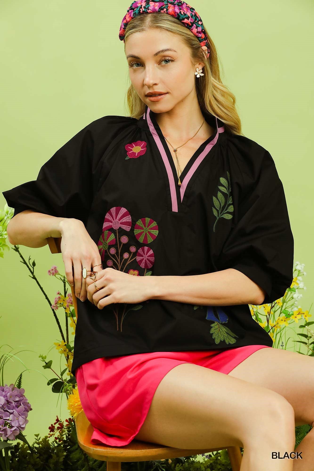 Woman wearing a black top with floral embroidery and pink skirt against a green background