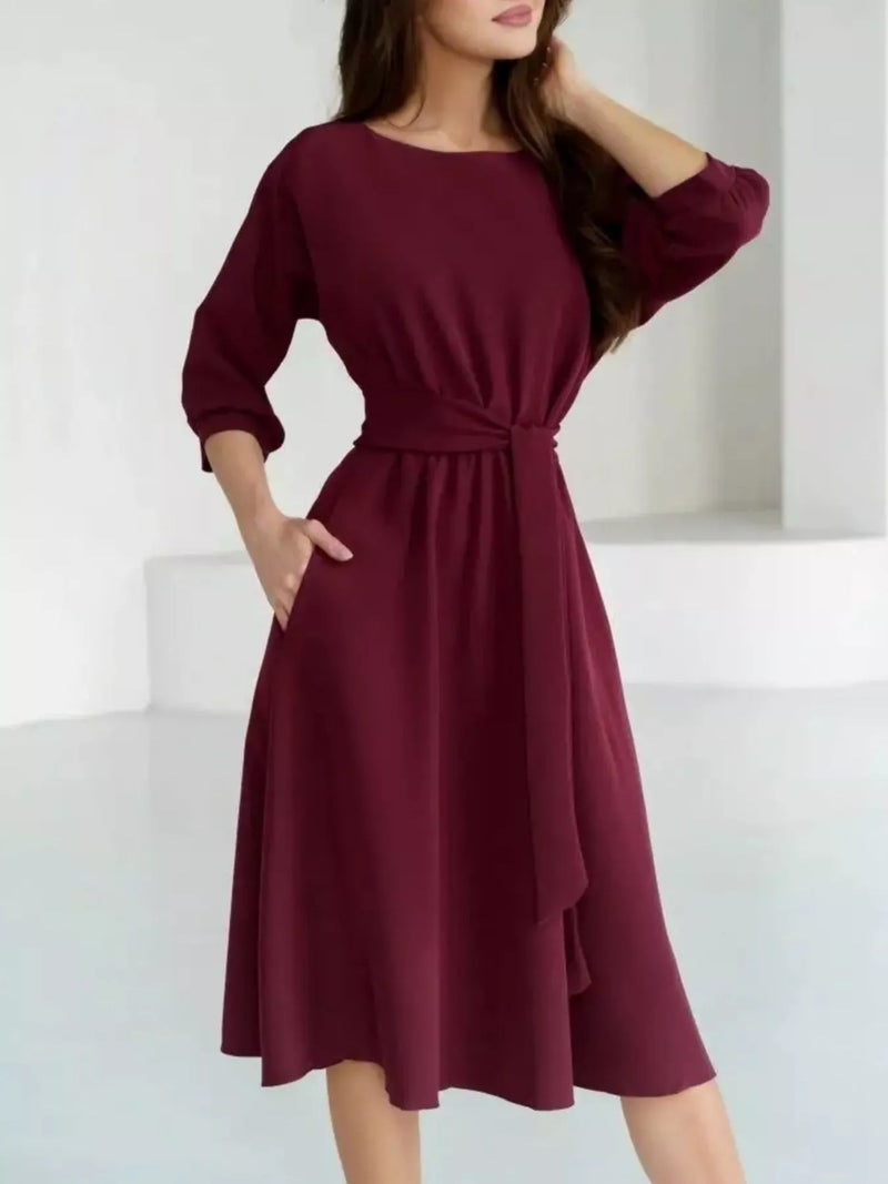 A woman wearing a wine-colored midi dress with three-quarter sleeves, a round neckline, and a tie-waist detail, set against a minimalist white background.