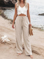 Woman in white top and beige pants standing on a sandy beach with water in the background.