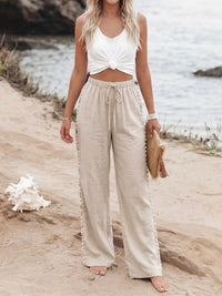 Woman in white top and beige pants standing on a sandy beach with water in the background.
