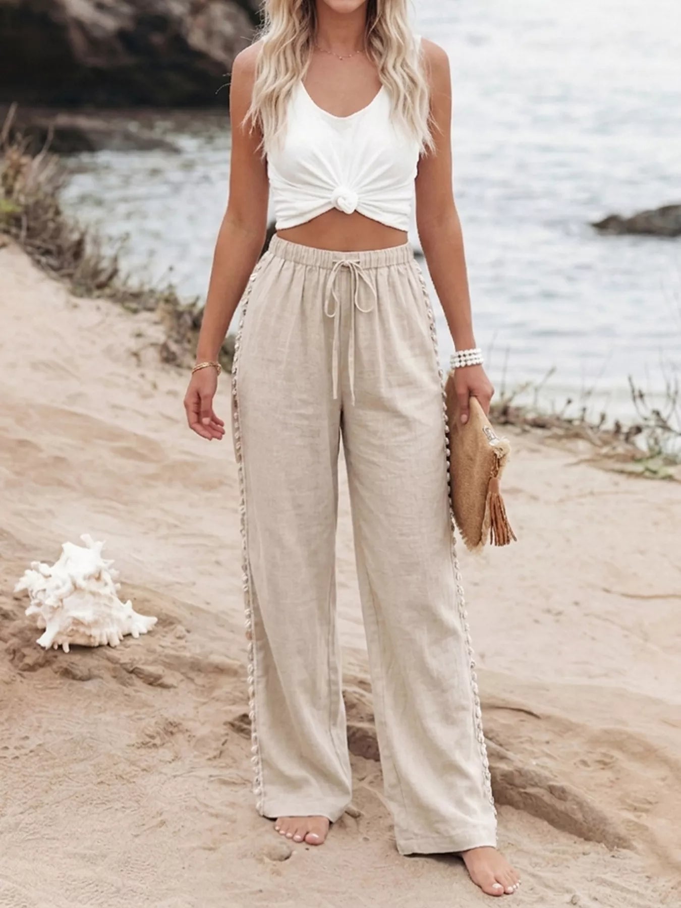 Woman in white top and beige pants standing on a sandy beach with water in the background.