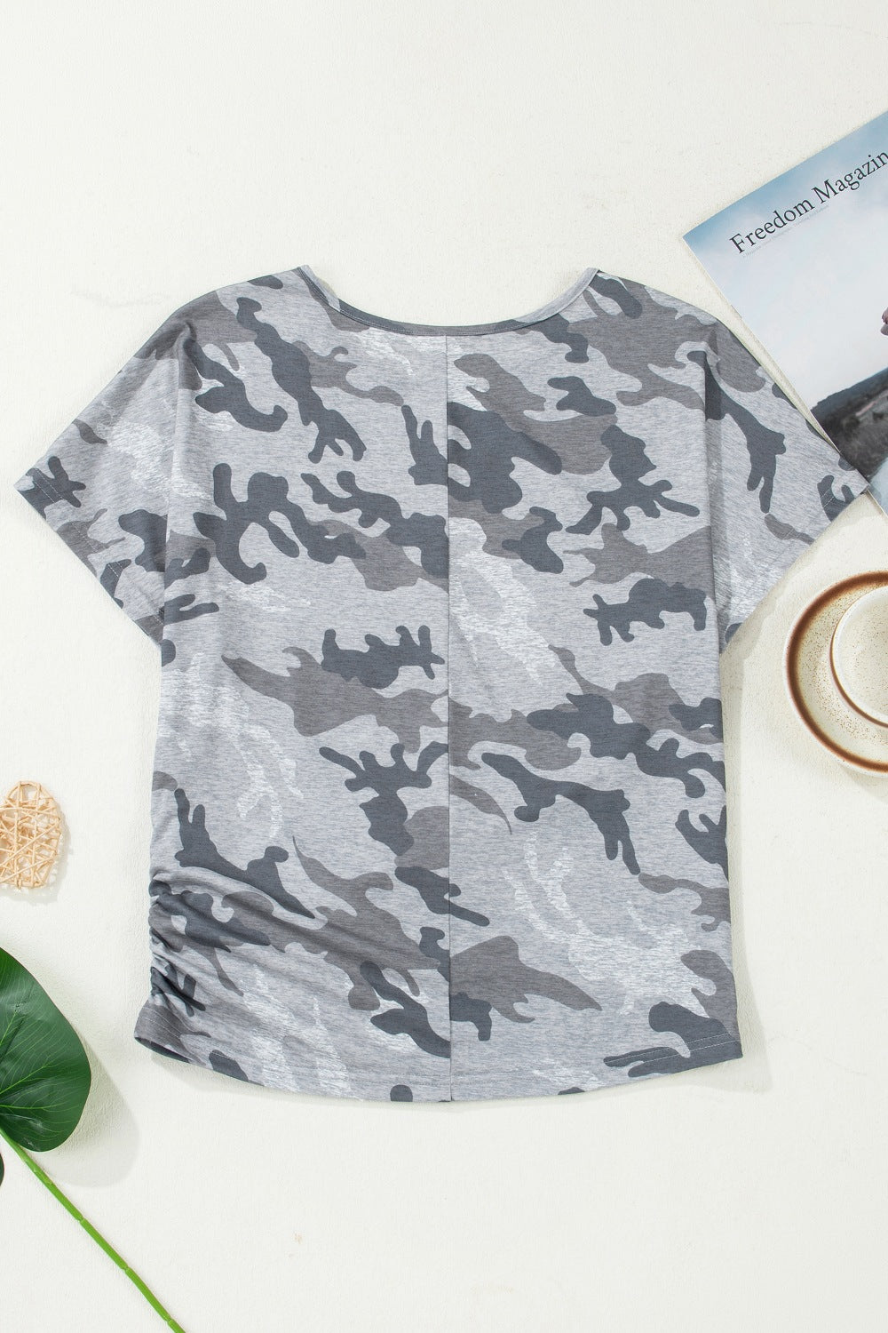 Camouflage-patterned shirt on a white surface with a magazine and cup in the background