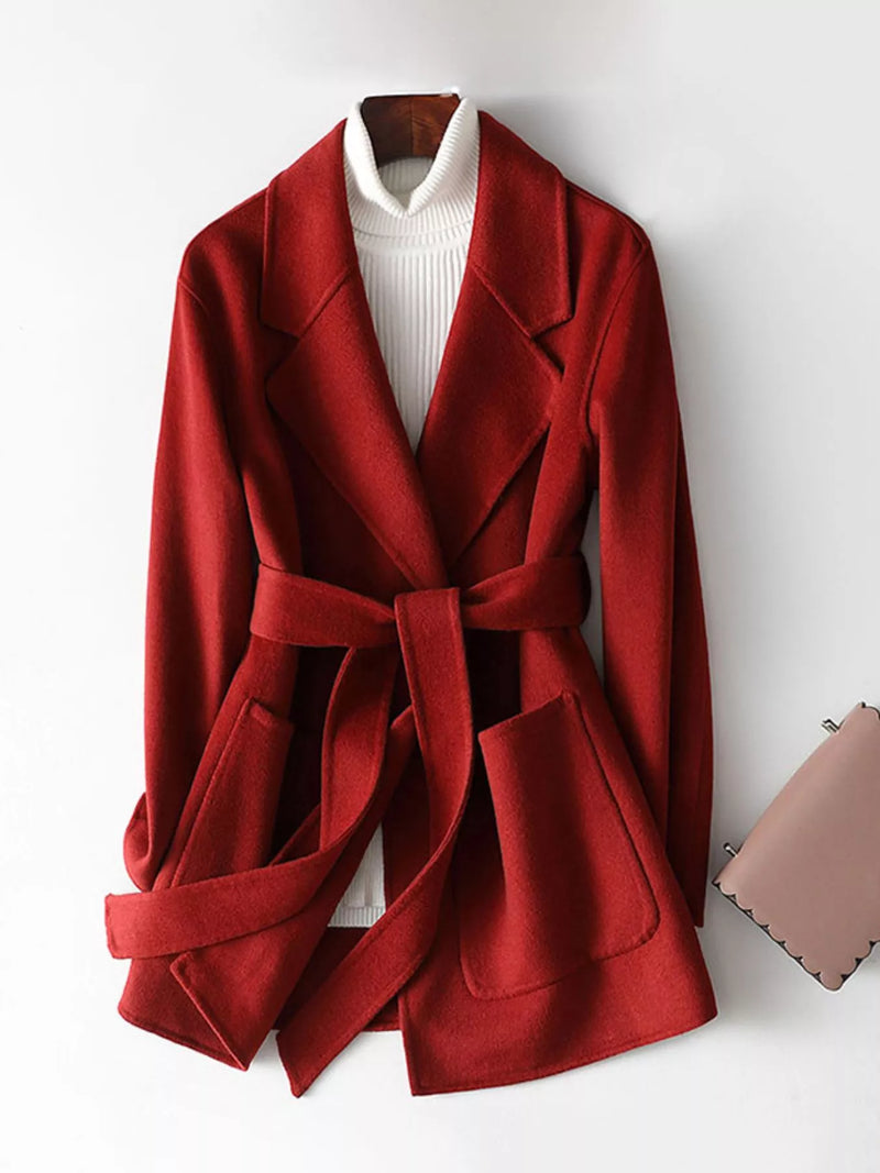 A red belted wrap coat with wide lapels, styled over a white ribbed turtleneck sweater. The coat is displayed on a hanger against a plain white wall, with a small pink purse partially visible on the right side.