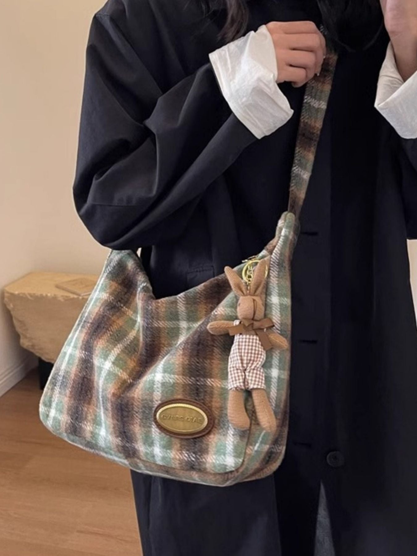 Plaid handbag with a teddy bear charm held by a person wearing a dark coat.