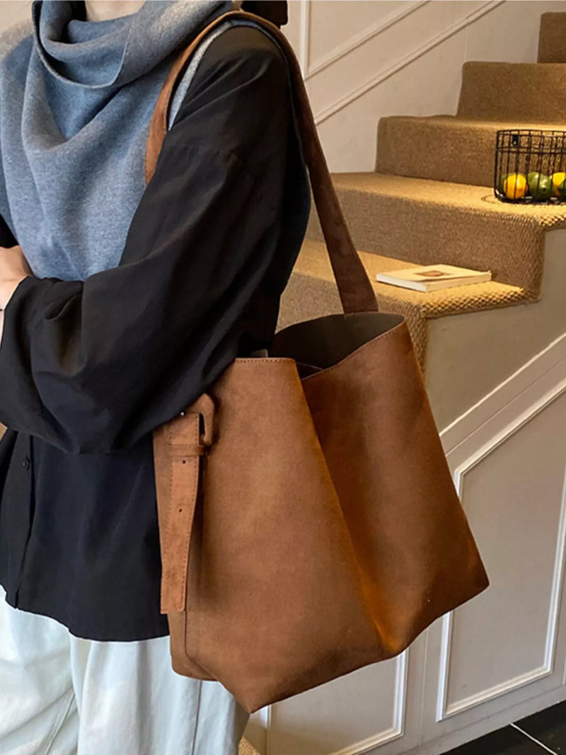 Person holding a brown leather tote bag in an indoor setting with stairs in the background.