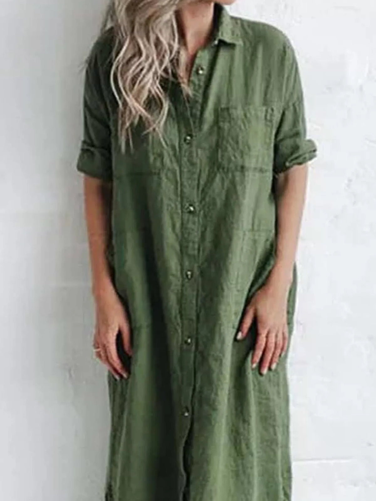 A woman wearing a long-sleeve olive green linen shirt dress with rolled-up sleeves and a button-down front, standing against a white textured wall.