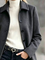 A close-up of a person wearing a dark charcoal gray wool-blend coat over a cream ribbed turtleneck sweater, paired with black jeans and a black belt with a silver buckle.