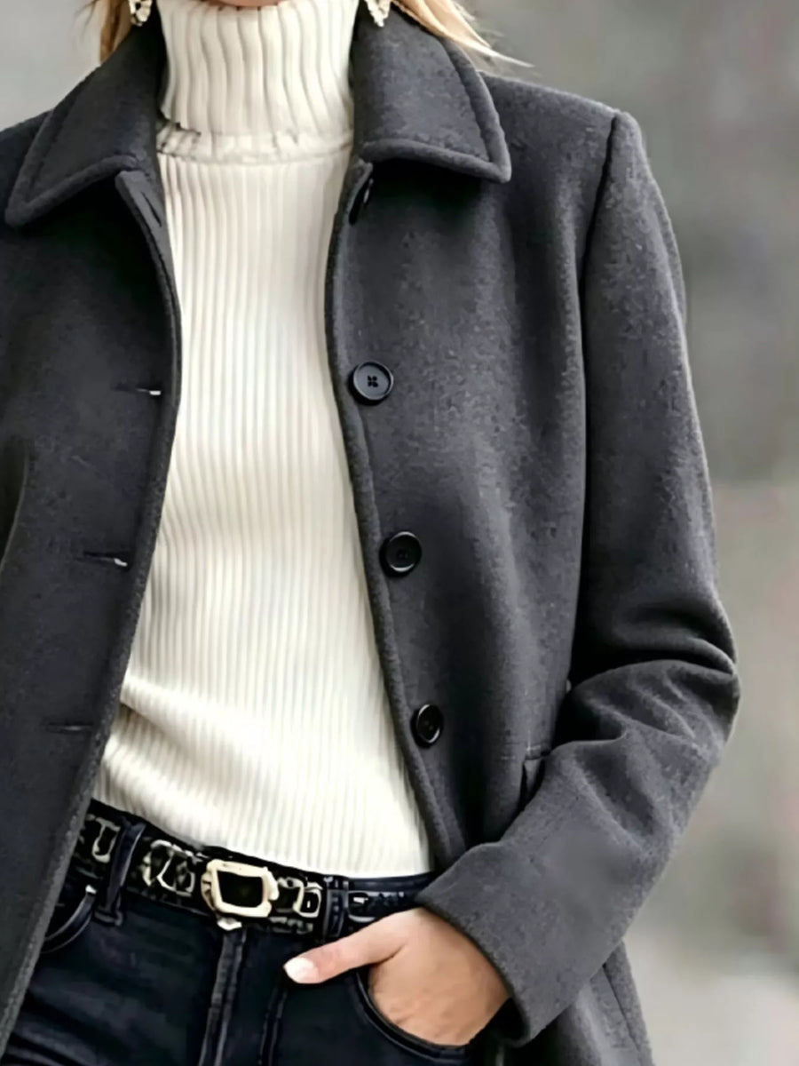 A close-up of a person wearing a dark charcoal gray wool-blend coat over a cream ribbed turtleneck sweater, paired with black jeans and a black belt with a silver buckle.