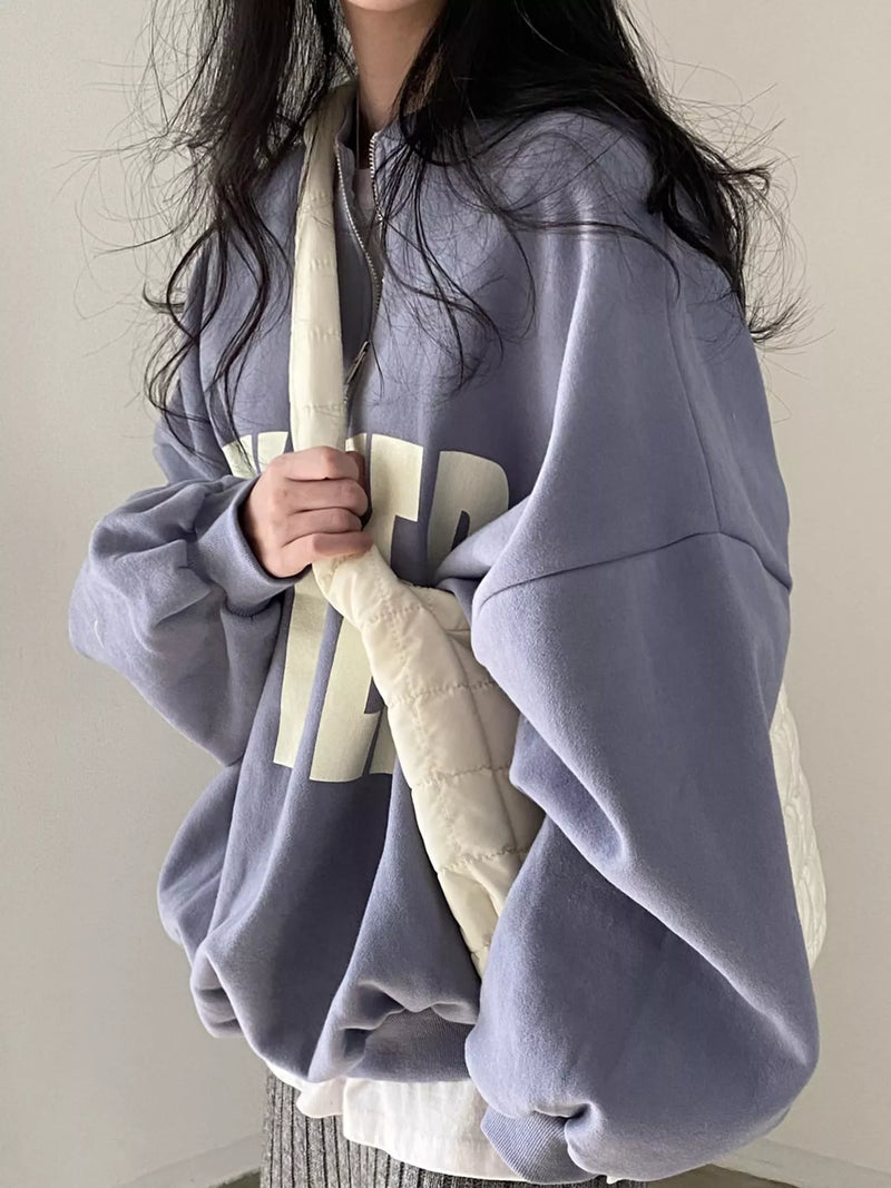A person wearing an oversized dusty lavender hoodie with a large cream graphic across the front, holding a cream quilted shoulder bag against a plain light background.