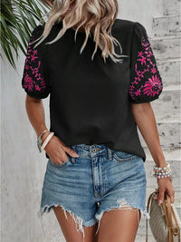 Woman wearing a black top with pink floral embroidery and denim shorts against a wooden wall.
