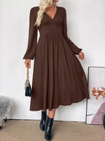 Woman wearing a brown dress with long sleeves against a white wall.