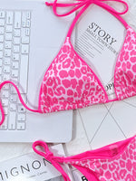 Pink leopard print bikini on a white surface with a laptop and magazine.