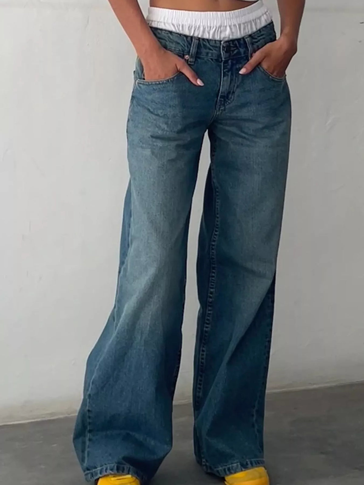A person wearing wide-leg blue denim jeans with a relaxed, baggy fit, paired with a white cropped top and yellow shoes, standing against a plain light gray wall.