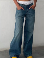 A person wearing wide-leg blue denim jeans with a relaxed, baggy fit, paired with a white cropped top and yellow shoes, standing against a plain light gray wall.